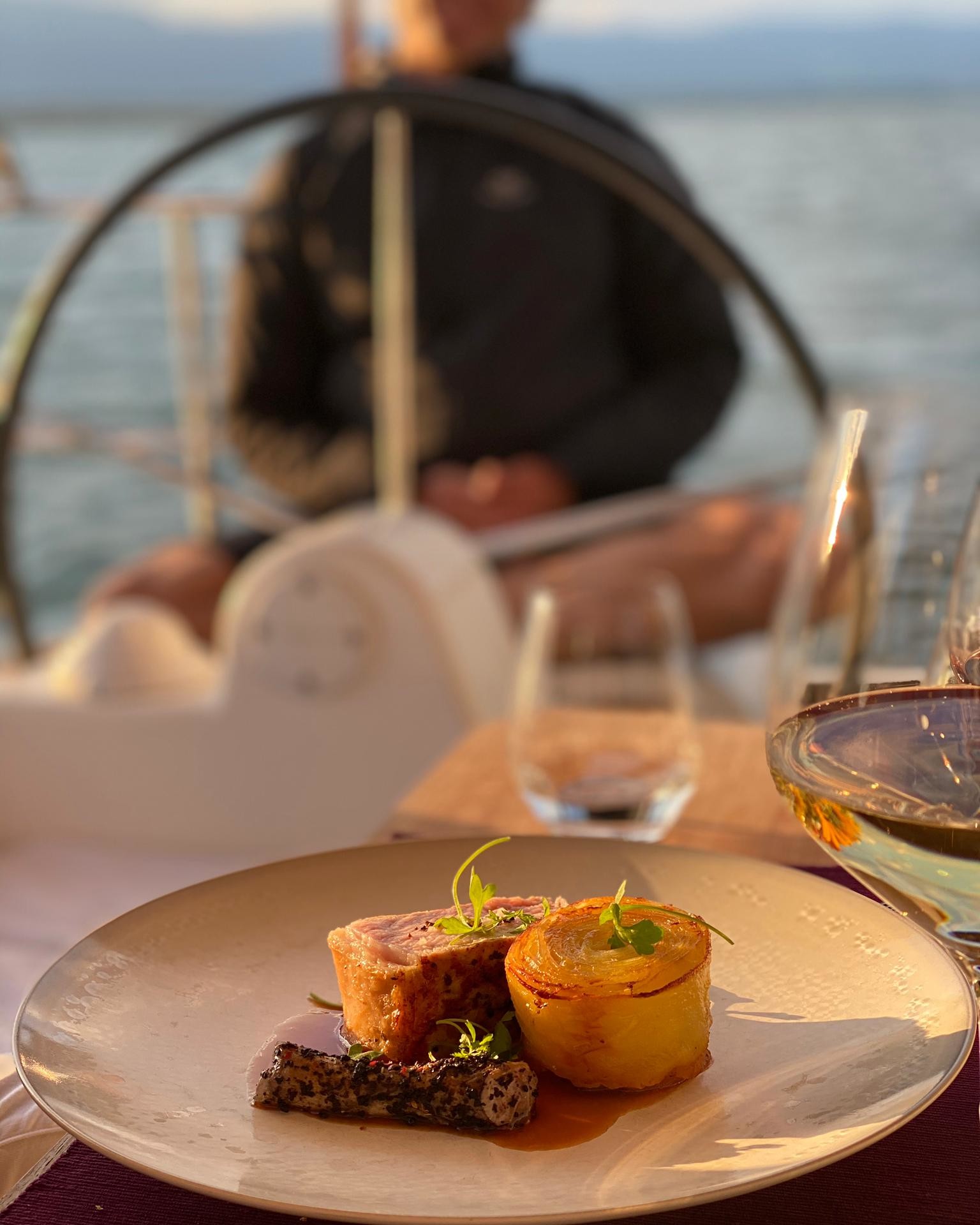 Plat gastronomique servi sur la terrasse de l’Hôtel Port Gitana à Genève, avec en arrière-plan le gouvernail d’un bateau sur le lac Léman : pièce de viande rosée, millefeuille de pommes de terre croustillant et sauce raffinée.