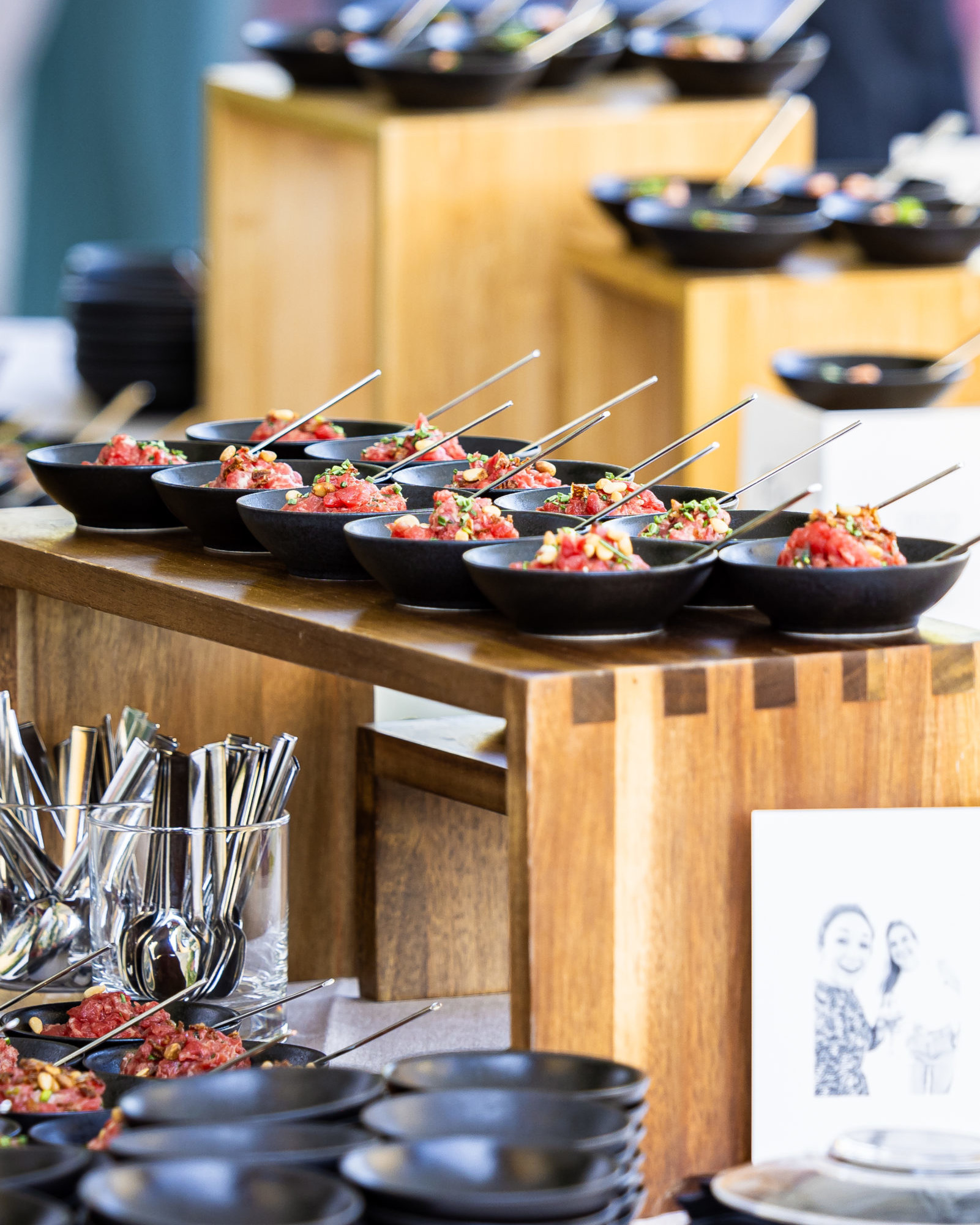 Buffet raffiné dressé dans un hôtel les pieds dans l’eau à Genève, mettant en avant une sélection de mets gastronomiques servis dans de petites assiettes noires élégantes. Les plats, joliment présentés avec des cuillères individuelles, sont disposés sur des présentoirs en bois naturel qui soulignent le côté chaleureux et convivial de l’établissement. Au premier plan, on aperçoit des piles d’assiettes et des verres remplis de couverts, prêts à accueillir les convives pour un moment gourmand au bord du lac Léman. Cette image illustre parfaitement l’expérience culinaire haut de gamme proposée par un hôtel les pieds dans l’eau à Genève, idéale pour un événement, un séminaire ou un séjour détente avec vue sur le lac.